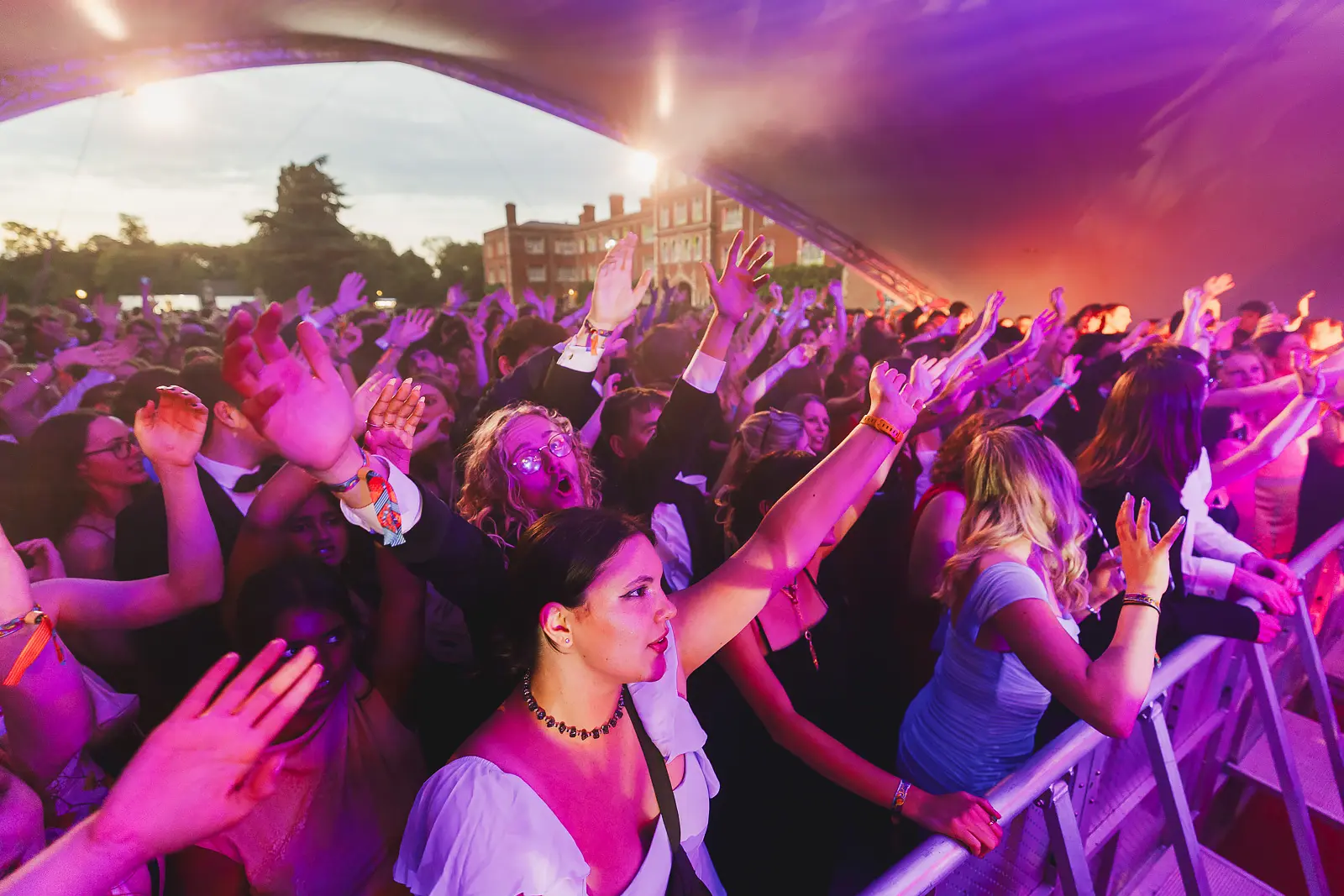Large crowd with arms raised under purple stage lighting at dusk with a college building visible in the background