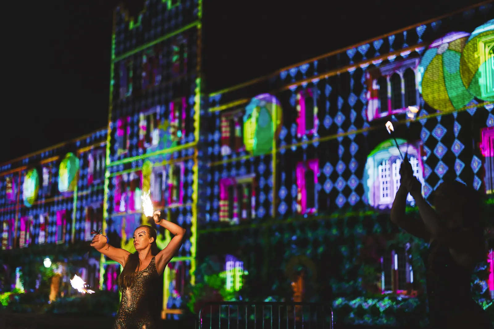 Fire dancer performing with poi flames in front of the neon projection-mapped college building at a summer ball at night