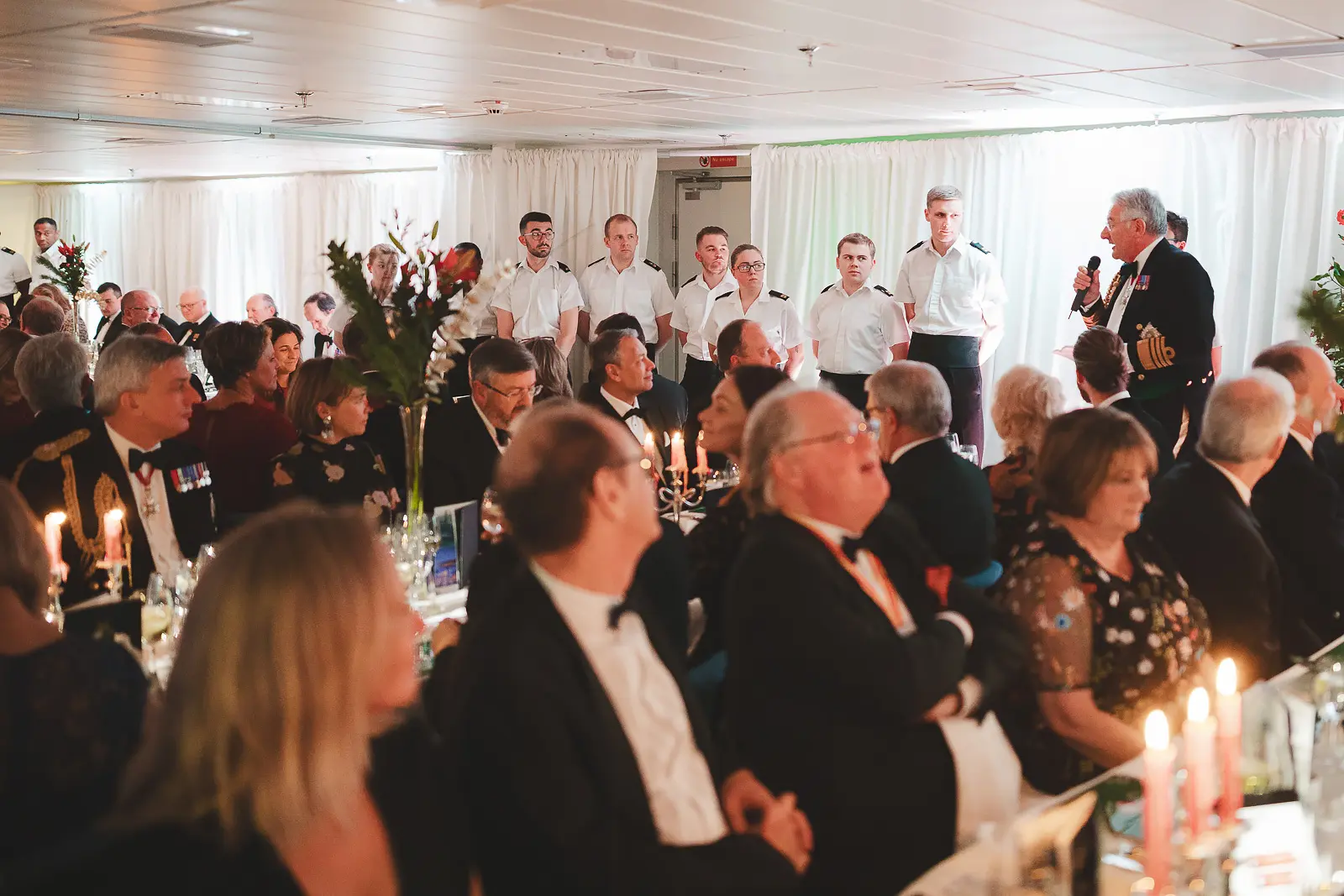 Wide view of formal black tie dinner in progress with candlelit tables, naval stewards in white at rear, officer delivering speech at microphone