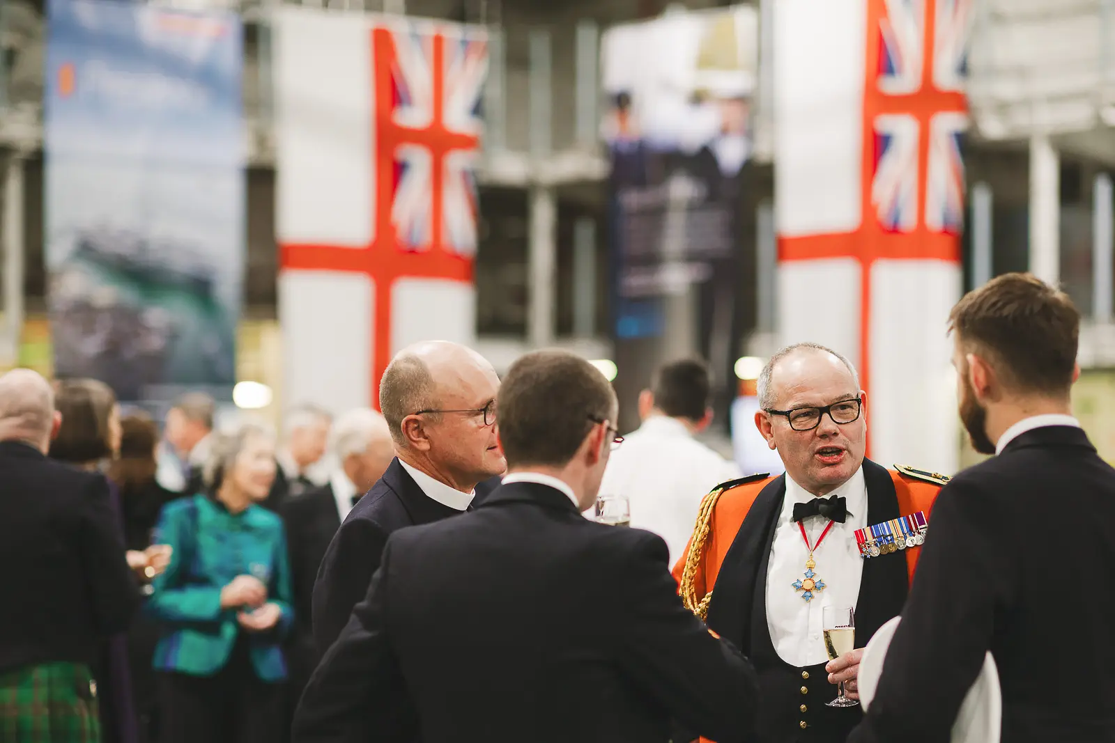 Guests mingling at formal drinks reception, senior officer in ceremonial dress with medals, Union flags in background
