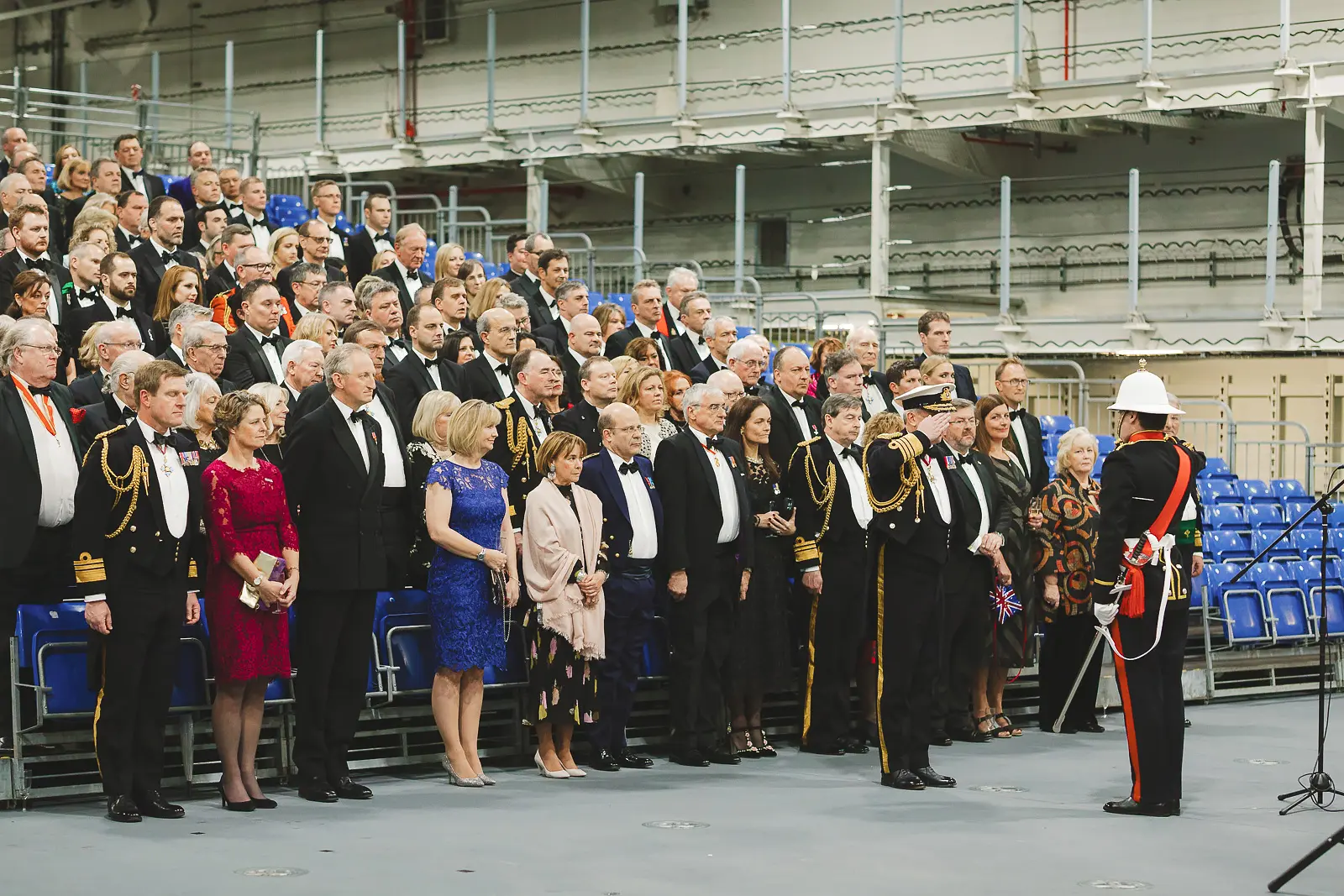 Large formal group of naval officers and black-tie guests assembled in rows for group photograph on hangar deck, drum major standing to one side