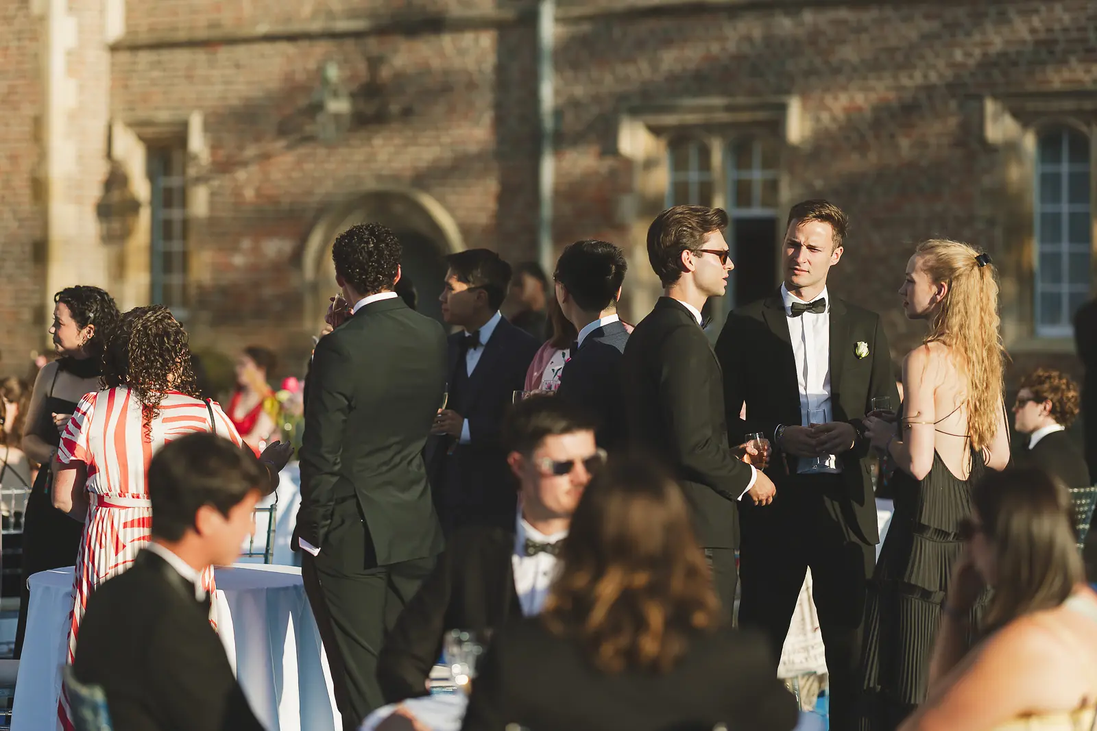 Formally dressed guests mingling in warm evening light against the brick facade of a historic Cambridge college