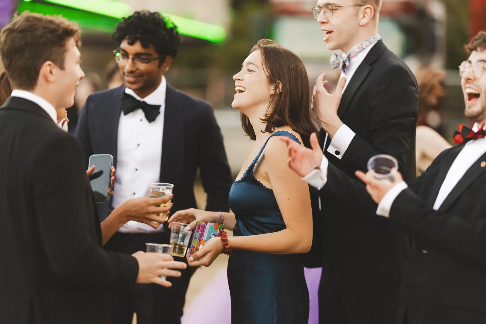 Group of formally dressed friends laughing together with drinks against neon fairground lights at a summer ball