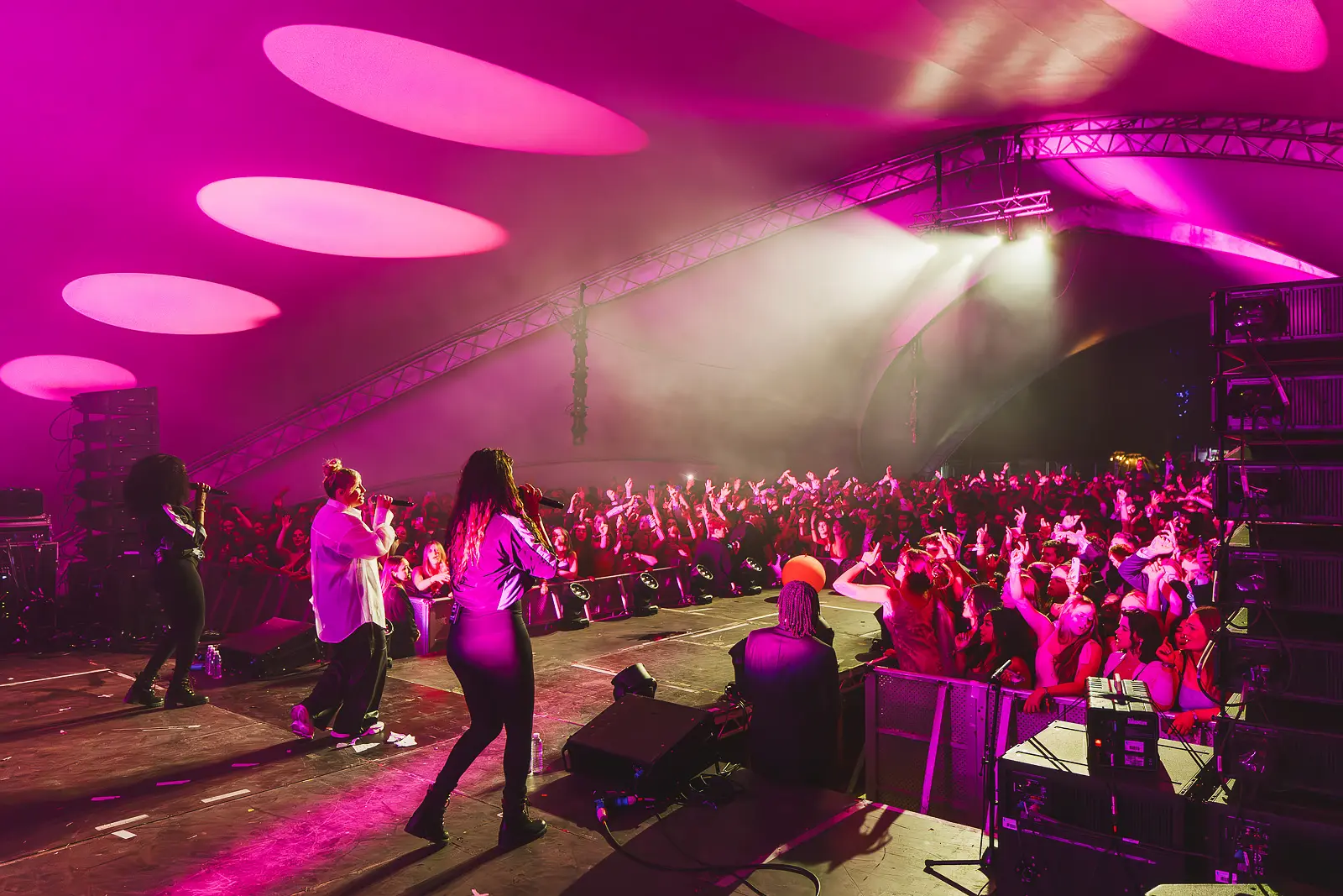 View from the stage across a packed tent venue with hands raised under pink spotlights at a Cambridge May Ball