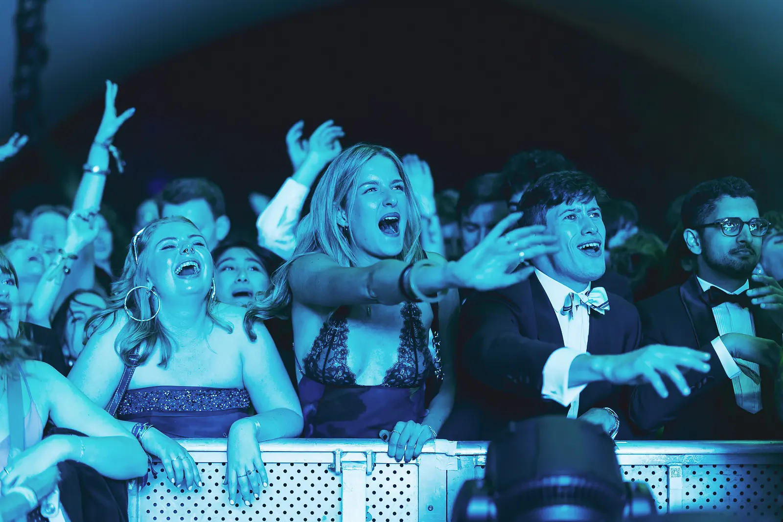 Guests in formal wear screaming and singing at the stage barrier under blue lighting during a summer ball concert