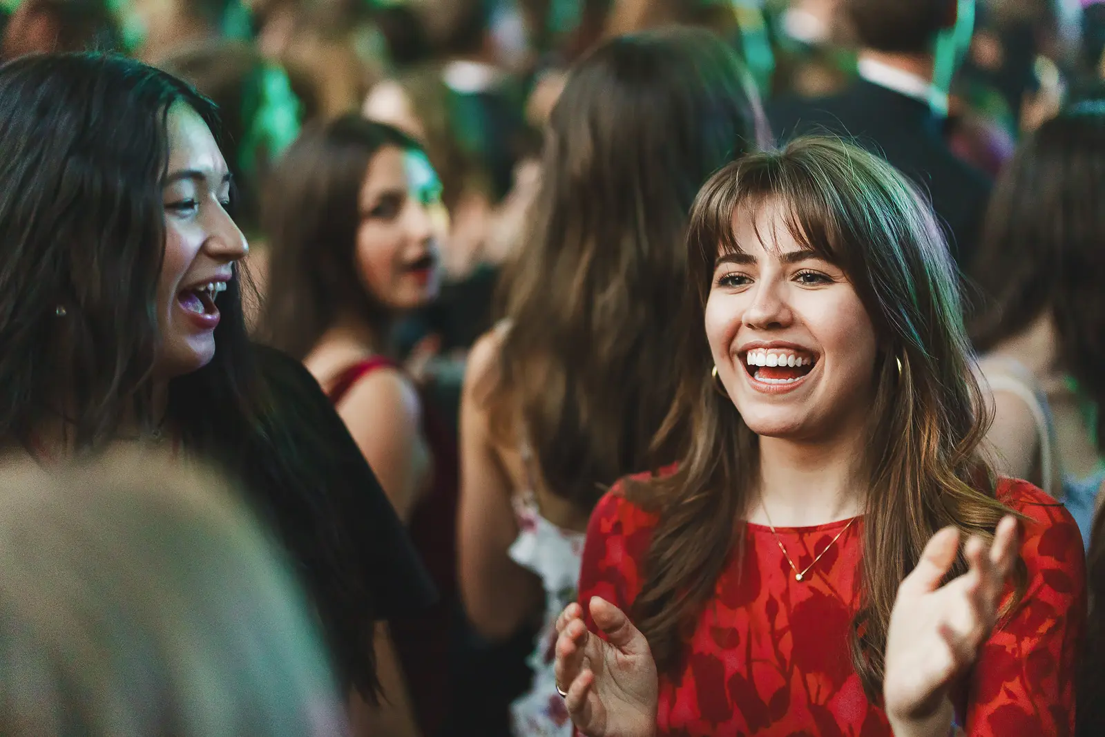 Two guests in evening wear caught laughing together in a crowd under green stage lighting at a summer ball