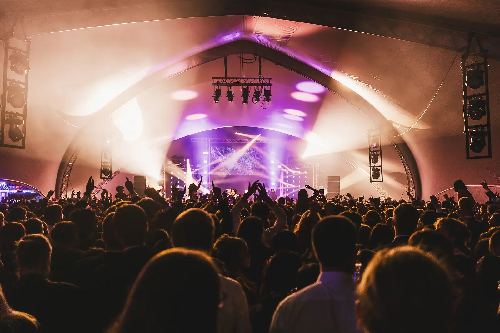 Crowd silhouettes with hands raised in warm amber and purple stage glow during a large-scale summer ball concert