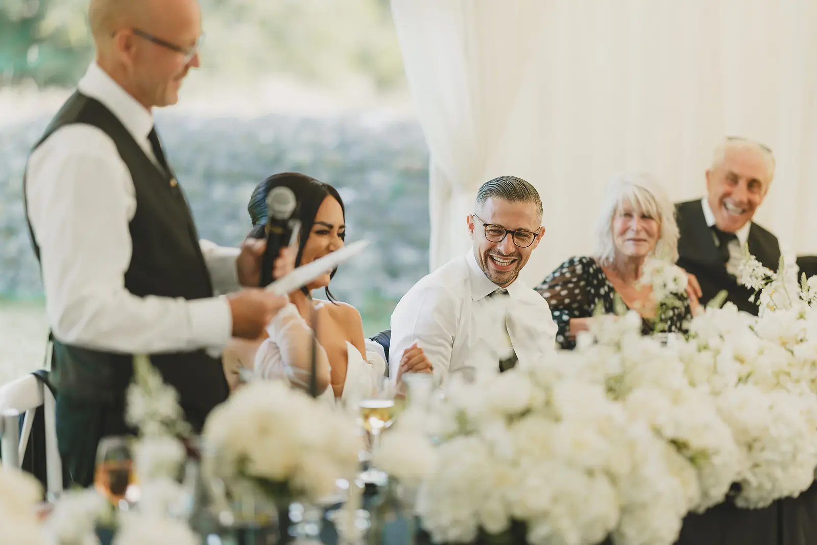 Groom laughing at the top table during the wedding speeches in the marquee, joyful reaction to the best man's words