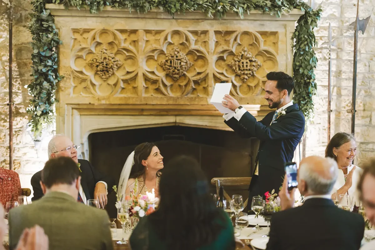 Groom reading his wedding speech notes as the bride looks up at him adoringly in front of the Gothic carved stone fireplace in the tithe barn