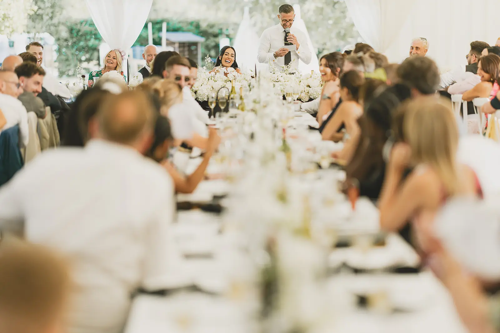 Groom delivering his speech to a full marquee of guests at the wedding reception, microphone in hand, guests attentive