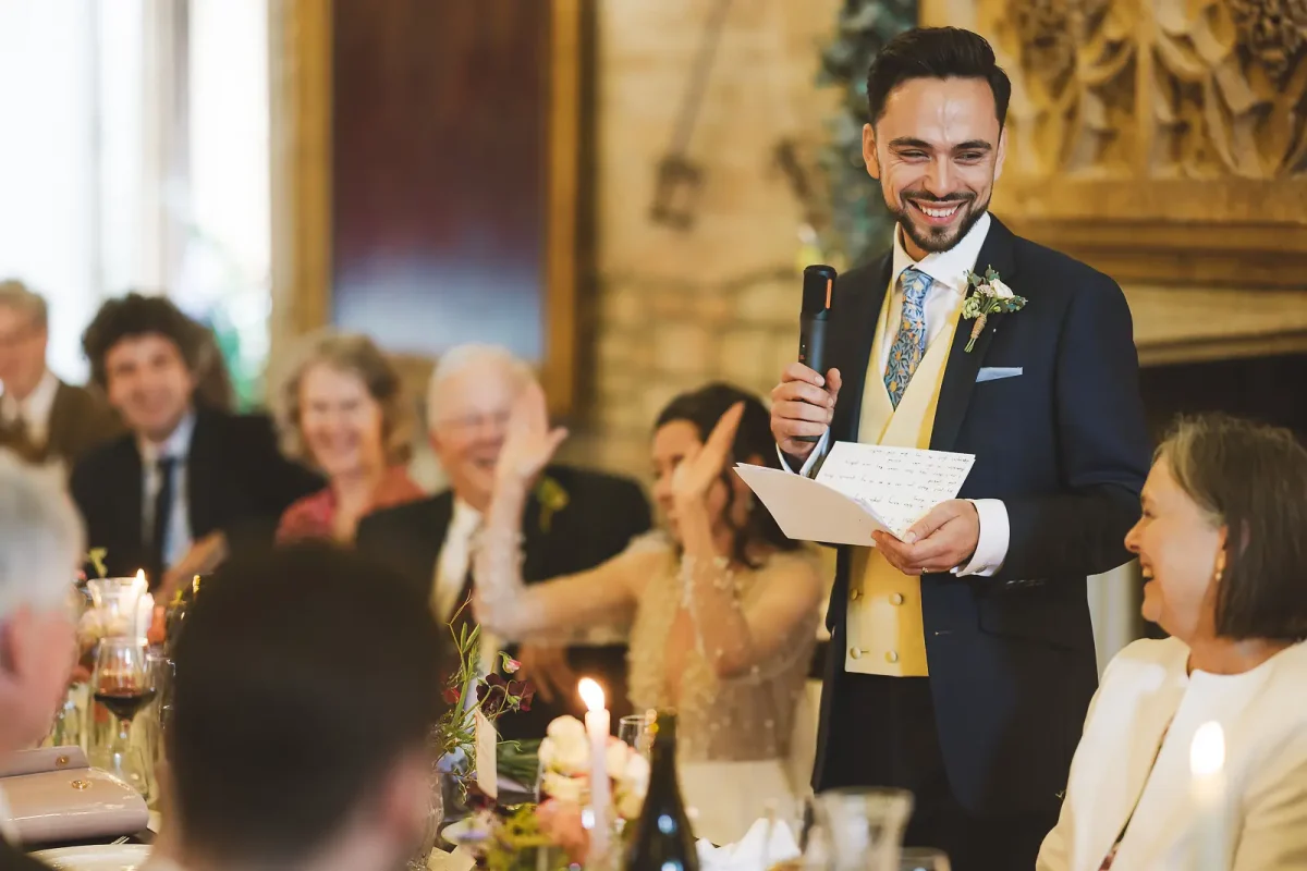 Groom smiling confidently as he delivers his wedding speech with guests laughing and clapping behind him beside the carved stone fireplace