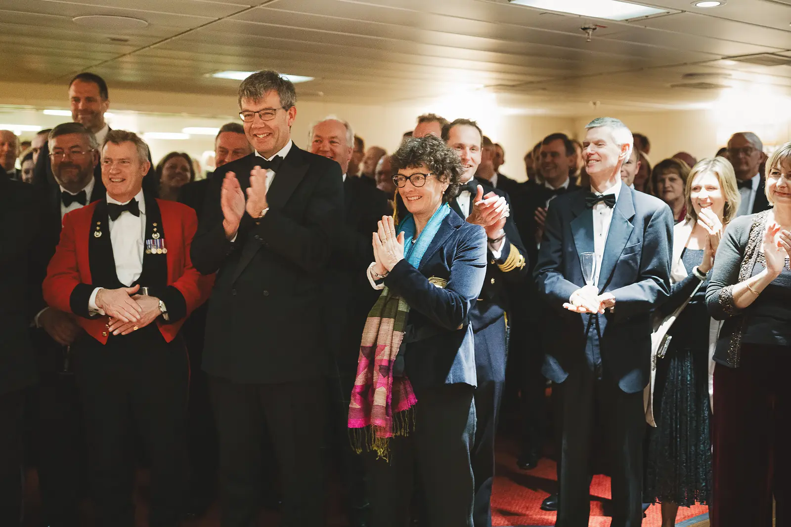 Mixed group of guests including officers in red mess dress and decorated dignitaries applauding at formal dinner inside ship's interior