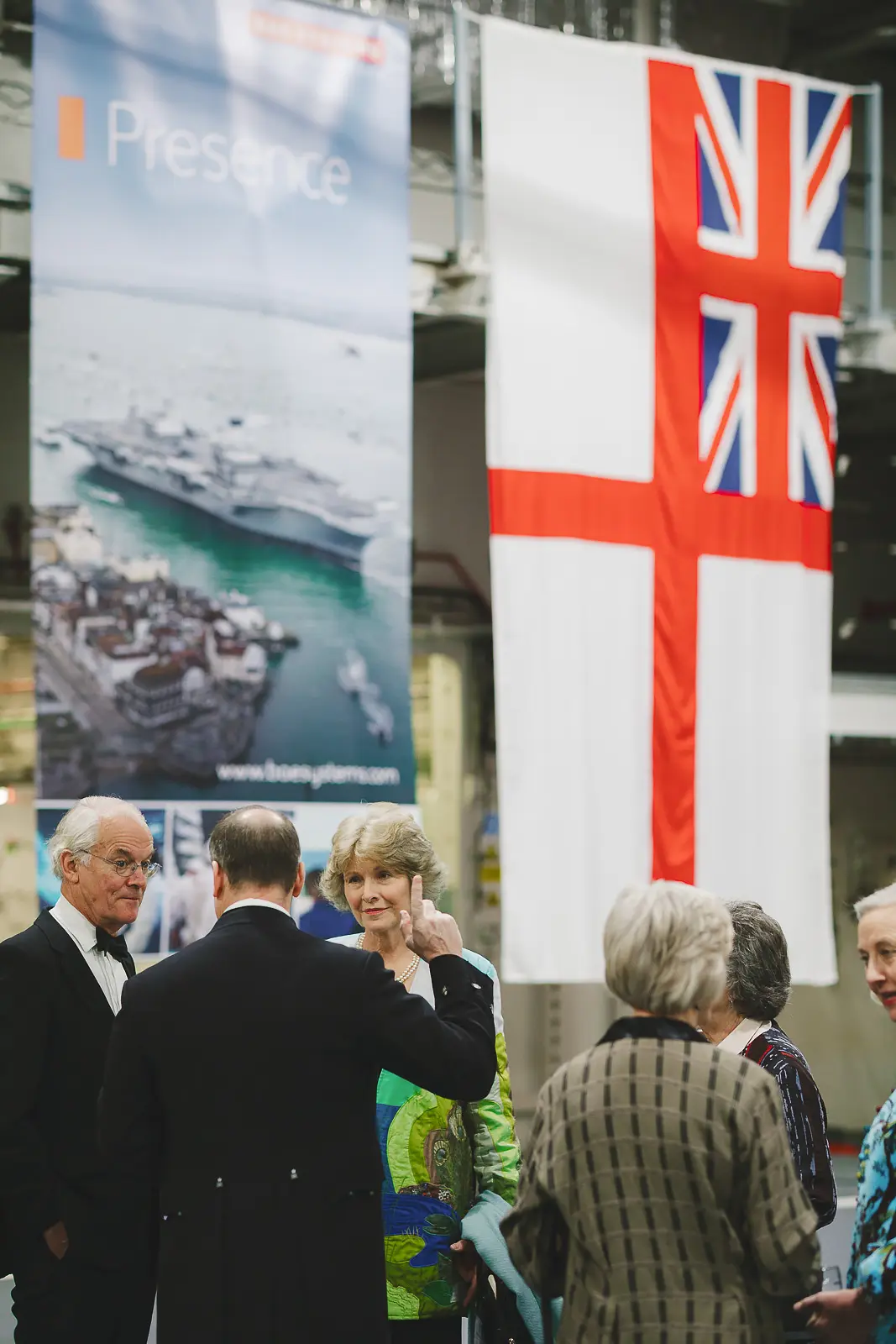 Guests in formal evening attire conversing beneath a large White Ensign and sponsor banner at military gala event, documentary photography