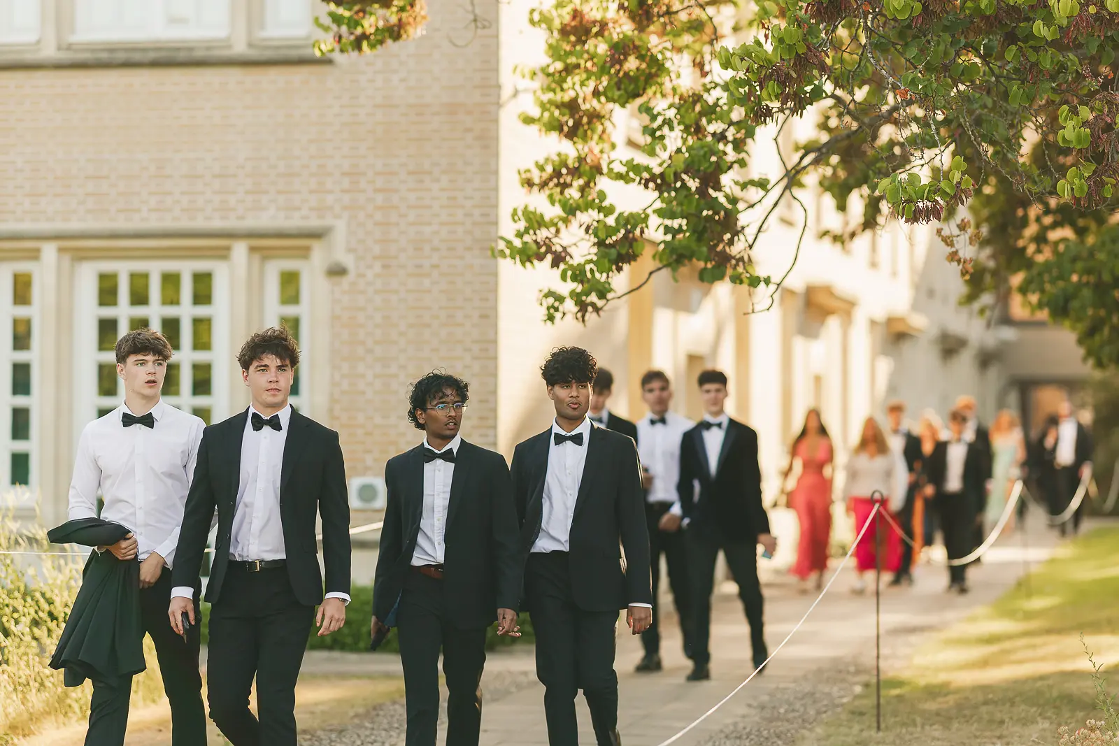 Young men in black tie walking along a college path in warm golden-hour light at a summer ball