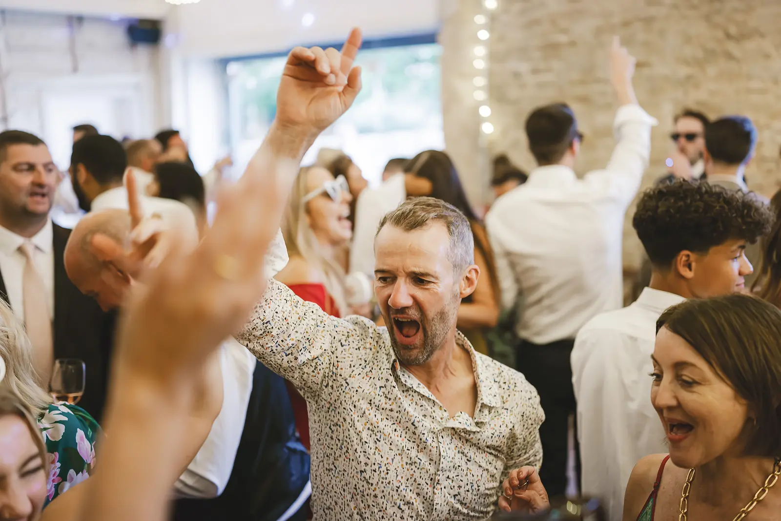 A crowd of wedding guests dancing together at the evening reception in the stone barn, fairy lights and warm atmosphere