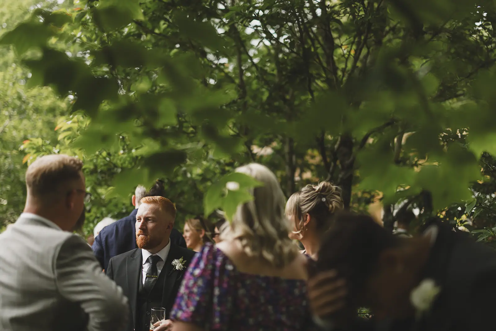 Wedding guests mingling and chatting under the shade of trees during the outdoor drinks reception at Mapperton
