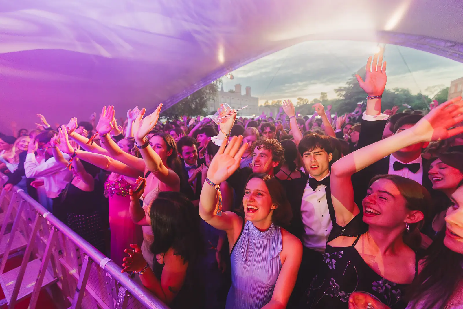 Crowd of formally dressed guests with hands raised under pink stage lighting at an outdoor summer ball concert