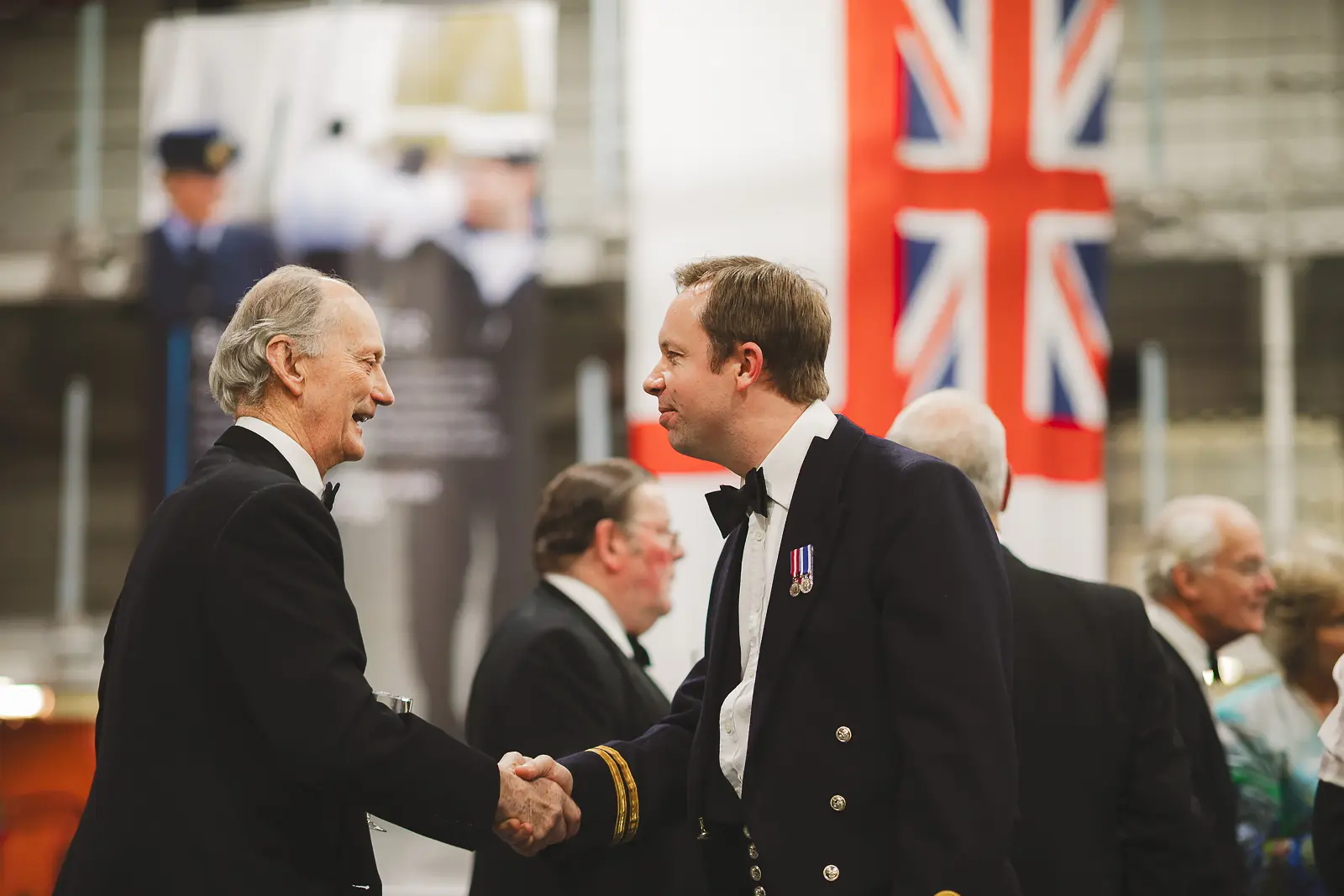 Naval officer with gold braid sleeve and medals shaking hands with civilian guest at formal gala, Union flag in background