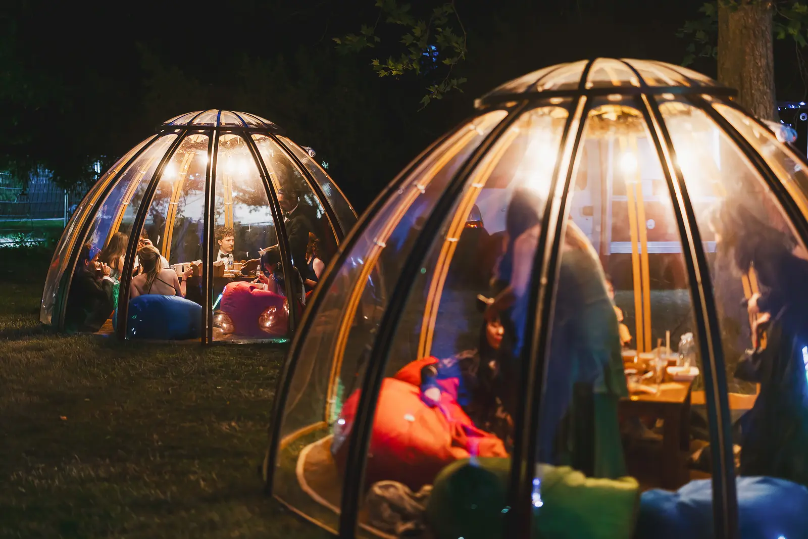 Illuminated transparent dome pods with formally dressed guests relaxing inside on bean bags at a May Ball at night