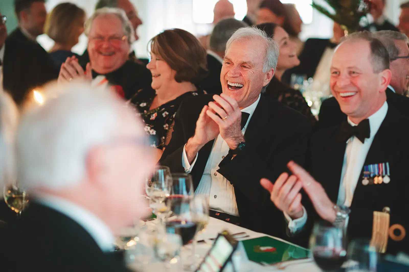 Joyful dinner guests laughing and clapping at formal gala dinner table, civilian and naval officer both mid-laughter, wine glasses in foreground