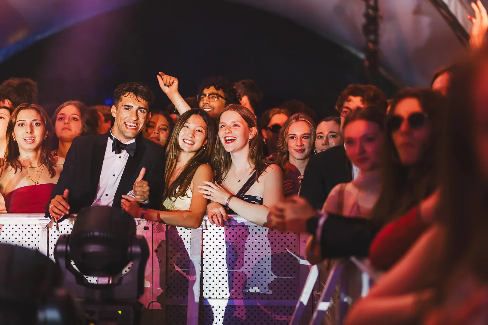 Smiling and cheering guests in formal wear pressed against the stage barrier at a May Ball concert