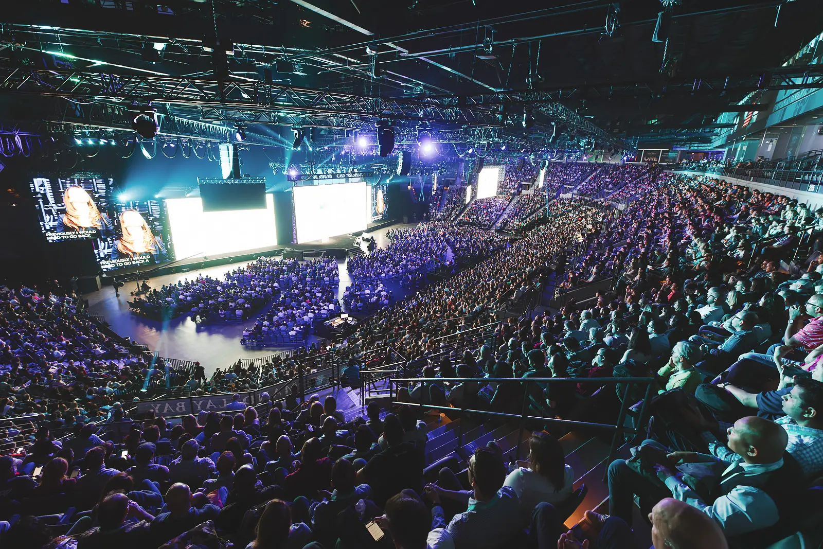 Large-scale conference audience filling an arena auditorium with stage lighting and screens
