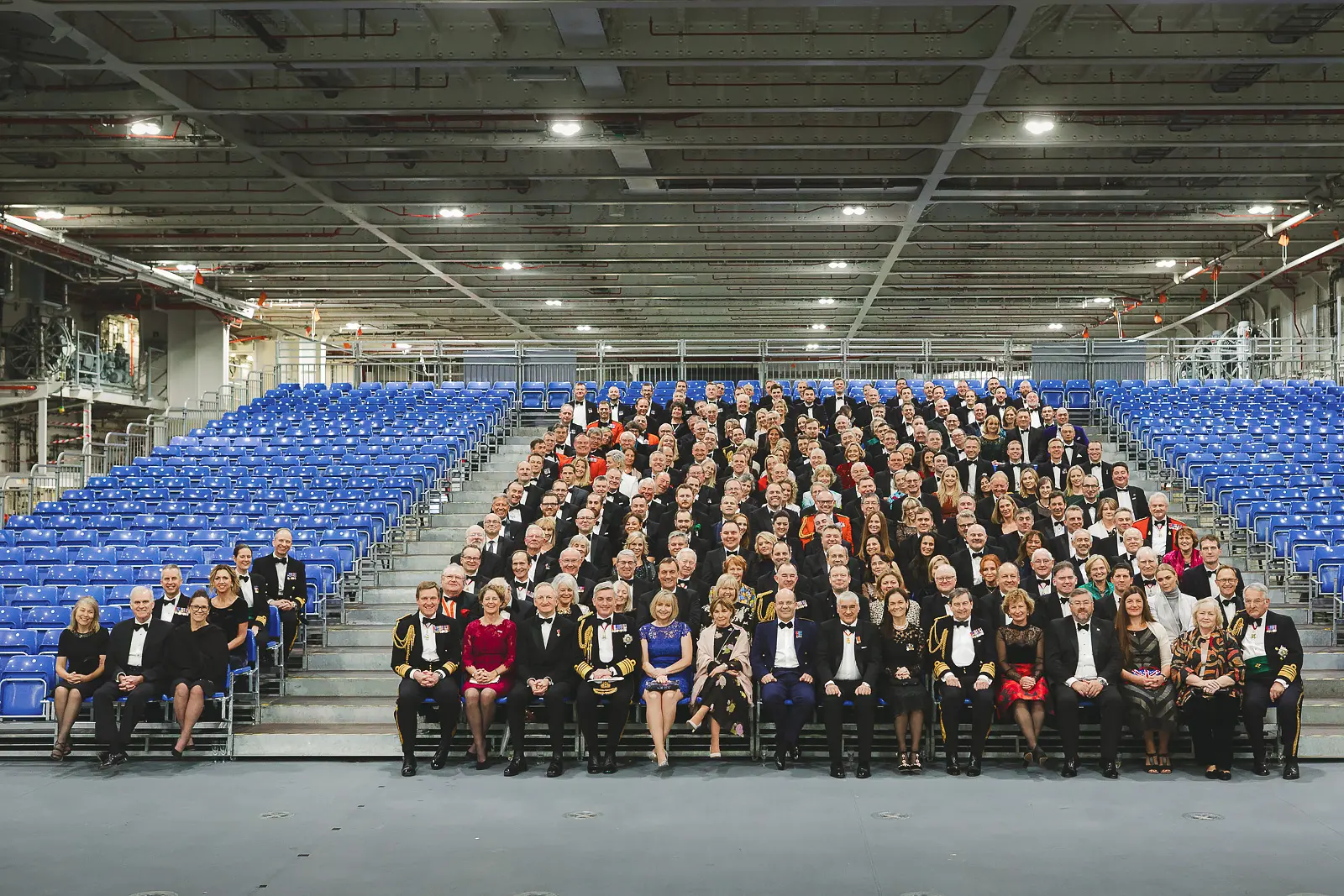 Formal group photograph of hundreds of guests in black tie and evening dress seated in tiered rows inside vast aircraft carrier hangar
