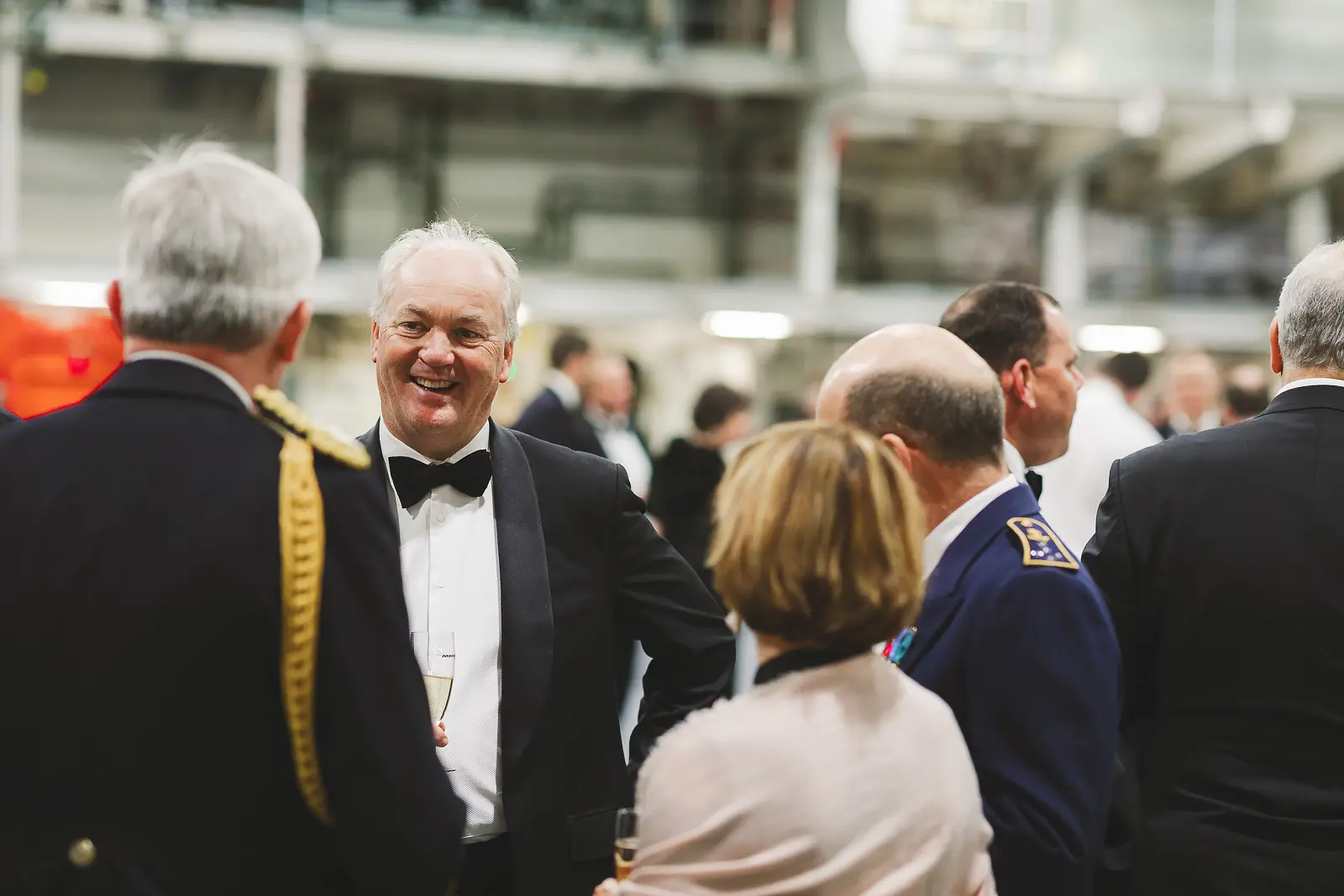 Smiling civilian guest in black tie laughing during evening reception on hangar deck, officers and guests surrounding him