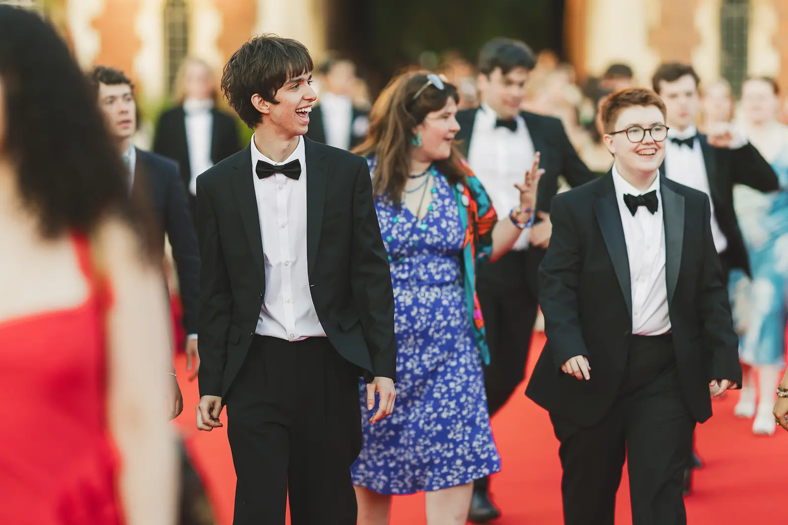 Young guests in black tie laughing as they walk along a red carpet entrance at a Cambridge May Ball