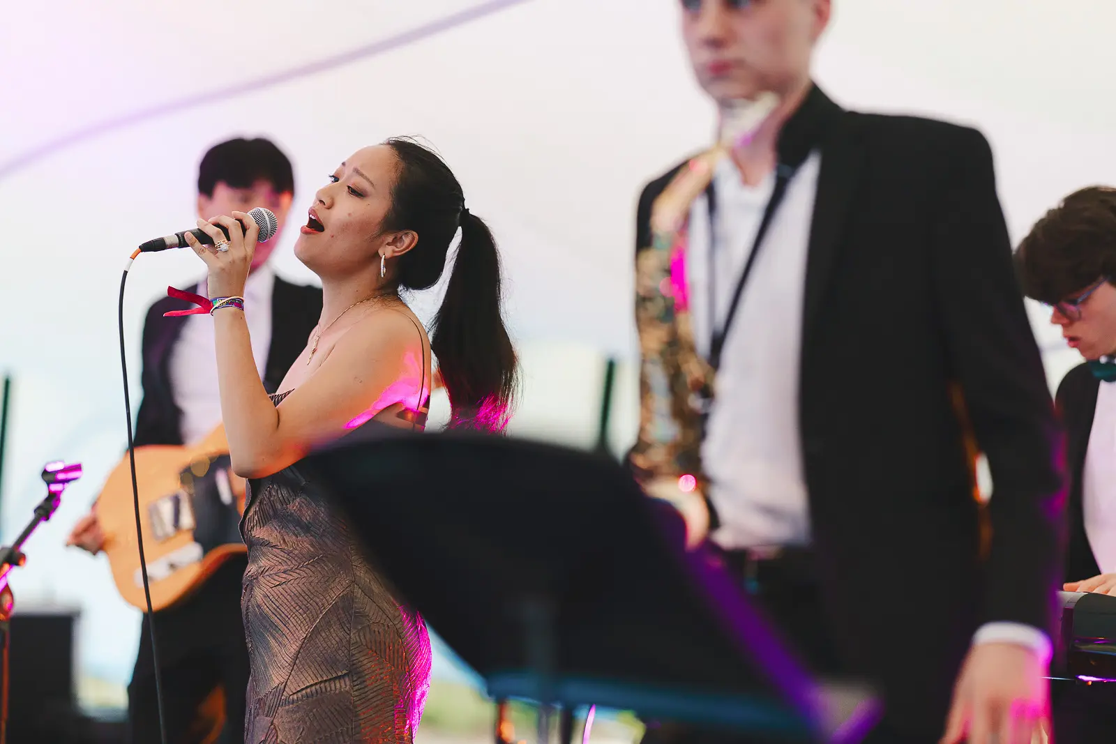 Female vocalist performing with a live band on stage under pink stage lighting at a Cambridge May Ball