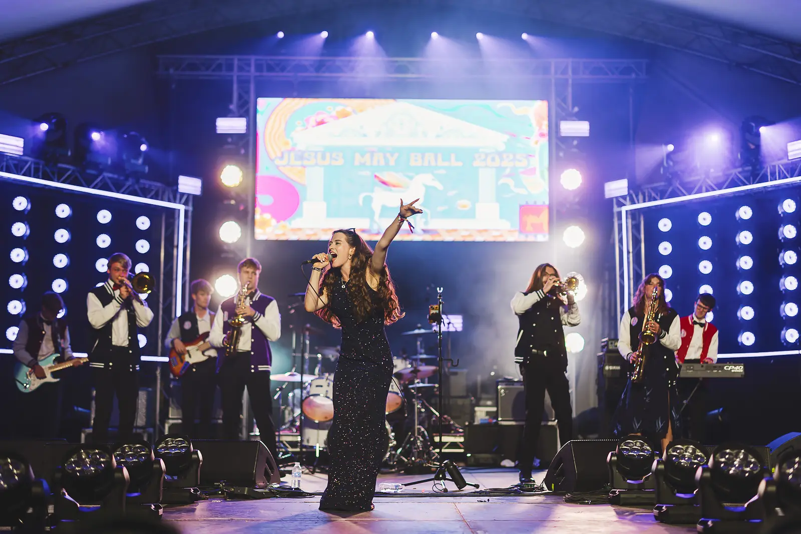 Female vocalist performing with arm raised on the Jesus May Ball 2025 main stage with a full band and brass section