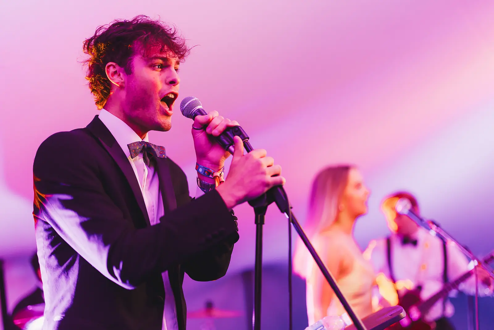 Male vocalist in black tie performing at the microphone under pink and purple stage lighting at a May Ball