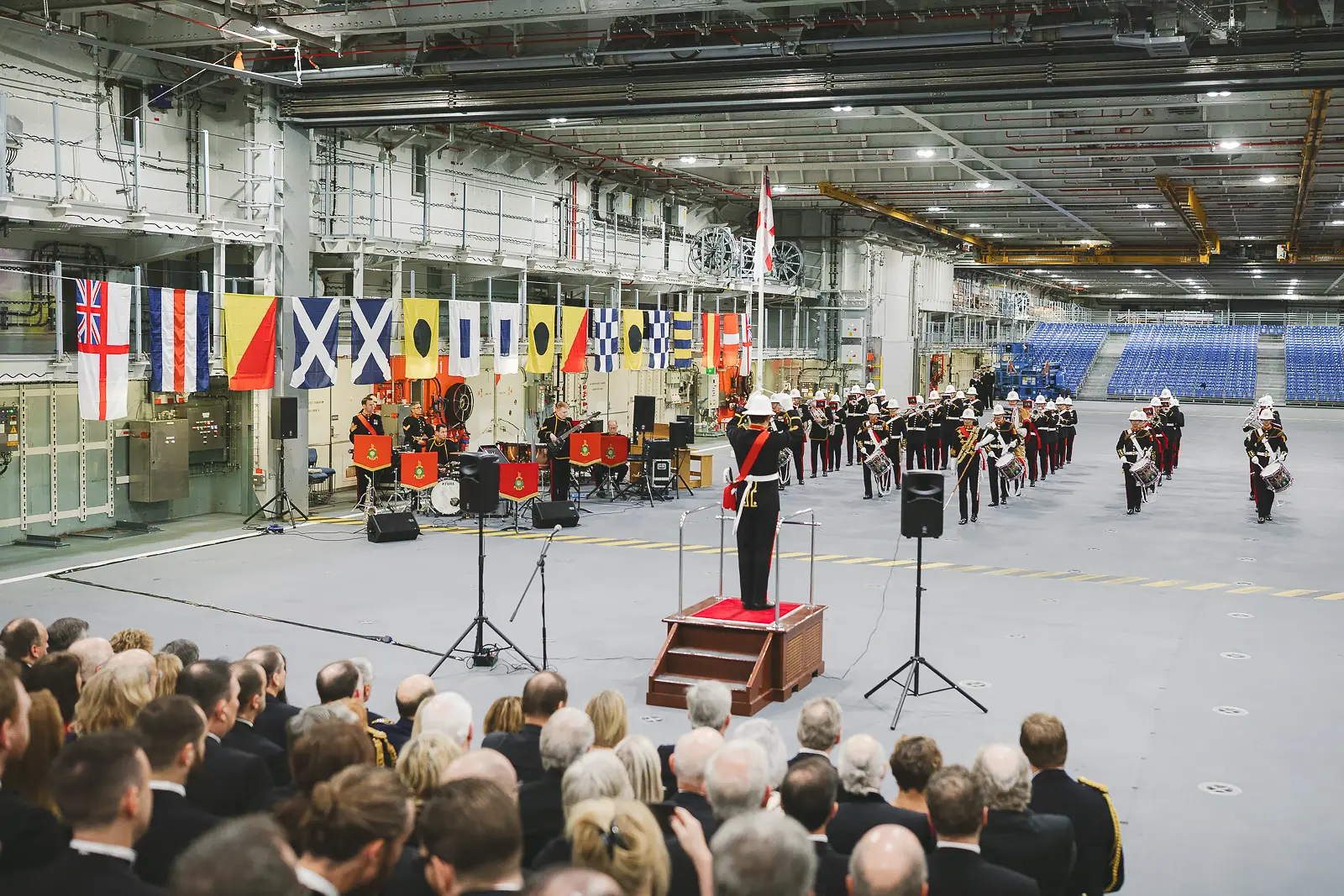 Royal Marines Band in ceremonial dress performing on hangar deck, naval signal flags along back wall, formal seated audience watching
