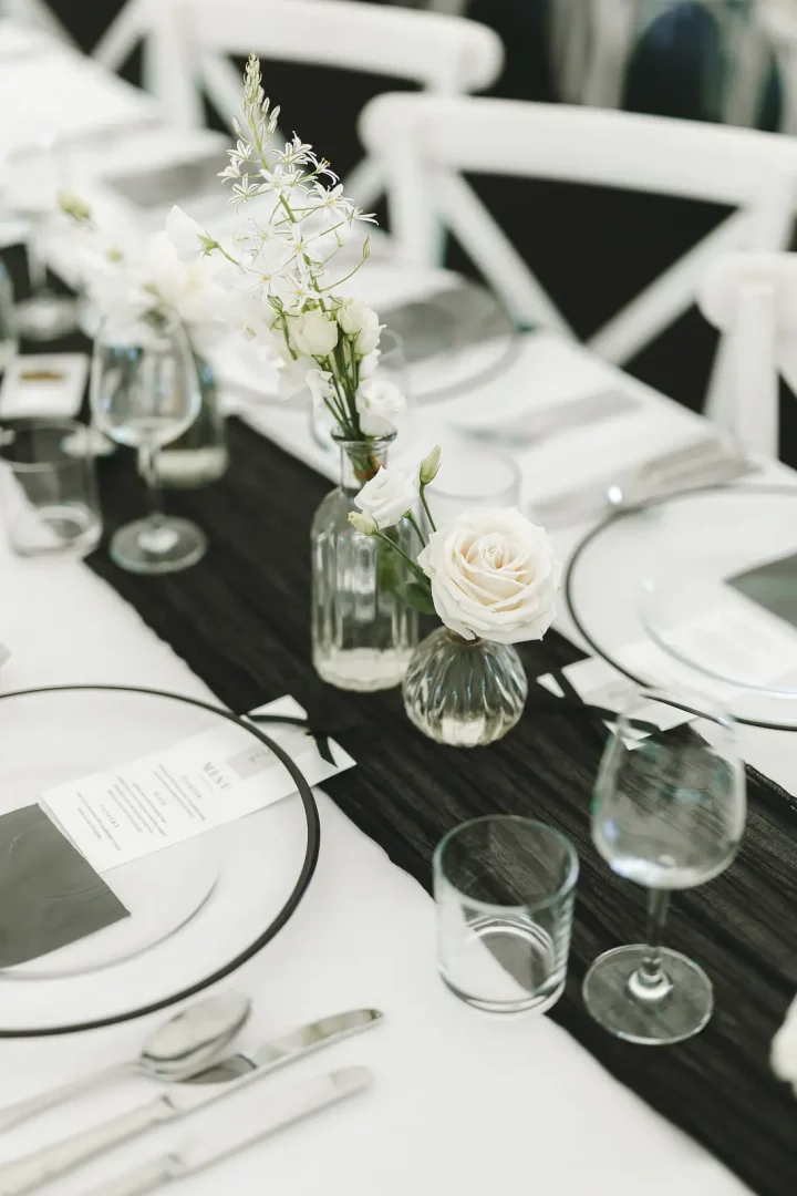Close-up of marquee table setting with black runner, white rose bud vases, rimmed plates, menu card and wine glasses, white cross-back chairs