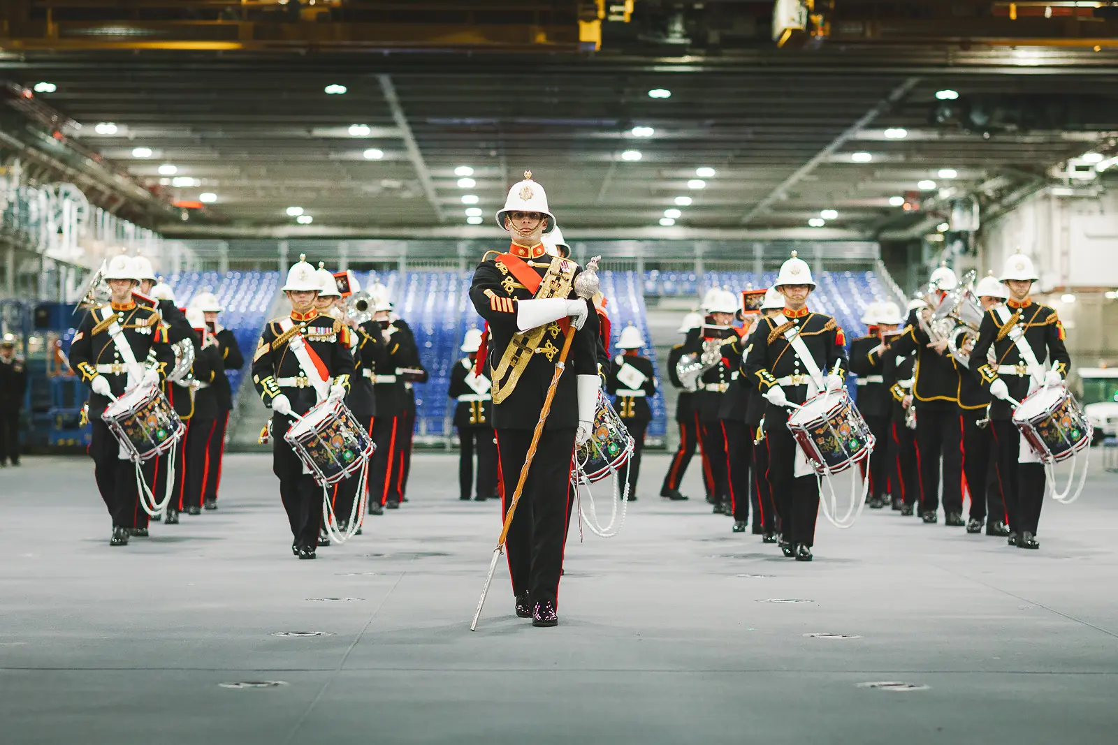 Military band in white pith helmets marching in formation on hangar deck, drum major leading at front with blue tiered seating behind