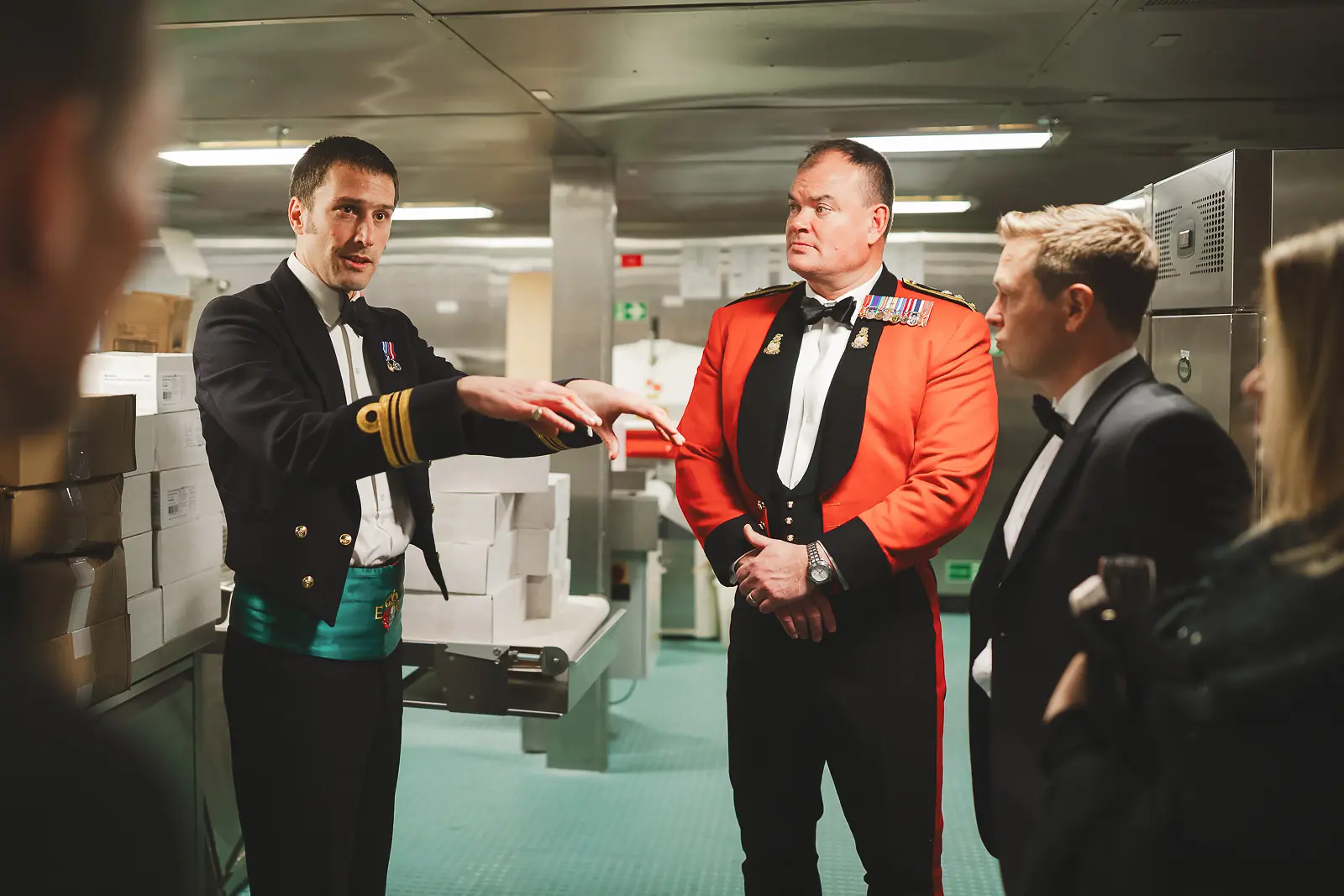 Naval officer gesturing to explain shipboard details to small group including officer in red mess dress and civilian guests during private tour