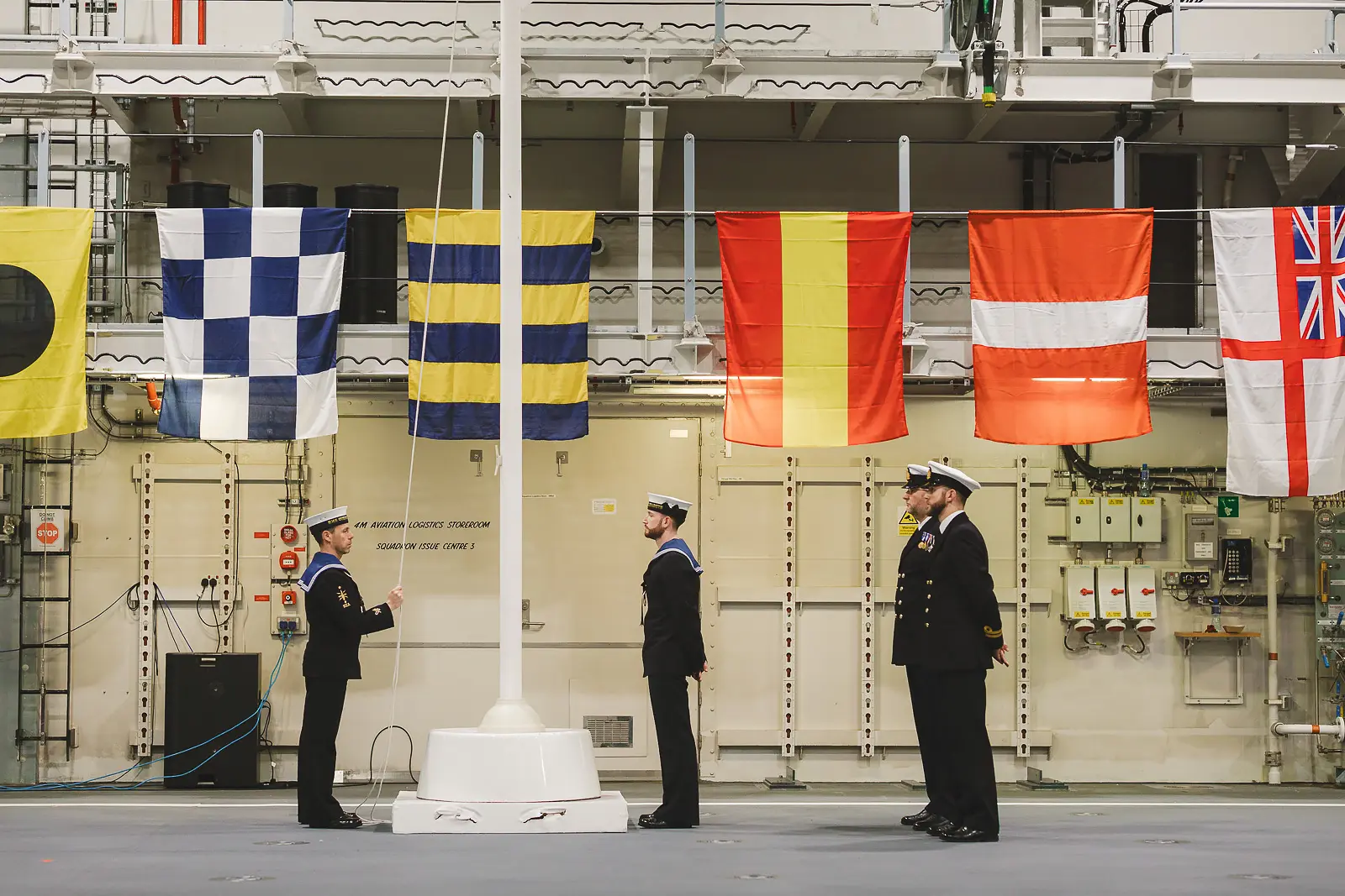 Three naval personnel standing at attention at mast base during formal flag ceremony, colourful signal flags and White Ensign displayed across hangar