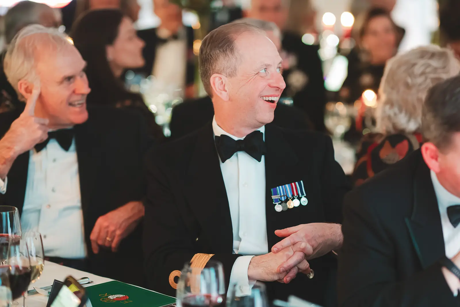 Naval officer with medals laughing and applauding at candlelit dinner table, civilian guest smiling beside him, candid reportage photography
