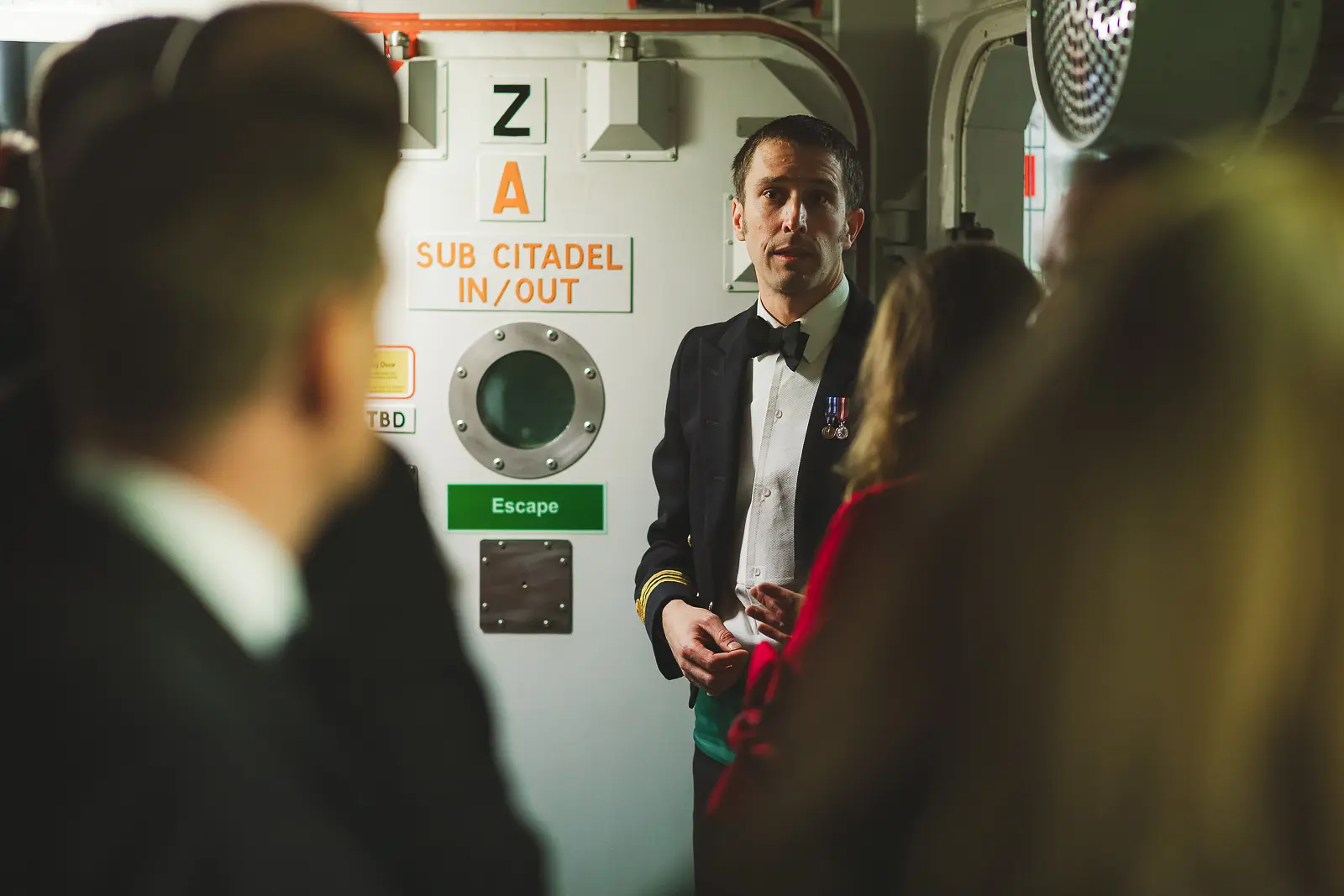 Naval officer in evening dress addressing guests beside Sub Citadel bulkhead door during private ship tour, escape signage and porthole behind