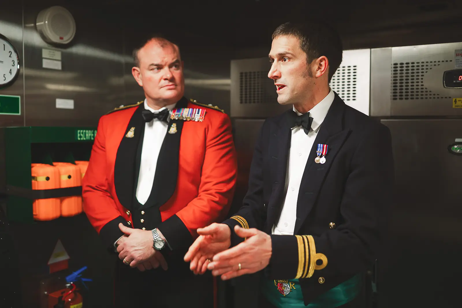 Officer in red ceremonial mess dress and naval officer in evening uniform during ship tour, life jackets and escape signage in background