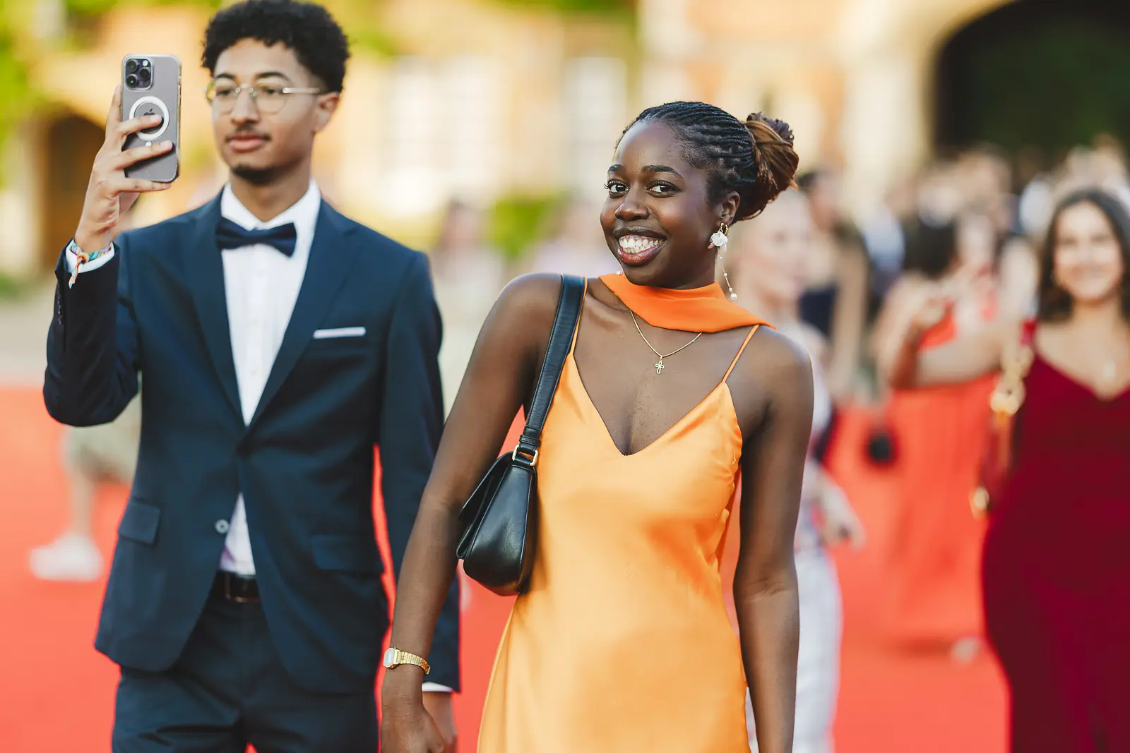 Guest in orange satin gown smiling on the red carpet as crowds arrive at a formal summer ball event