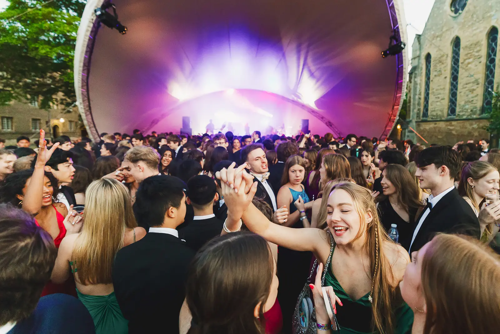 Dense crowd of formally dressed guests dancing in front of an outdoor main stage at a Cambridge May Ball