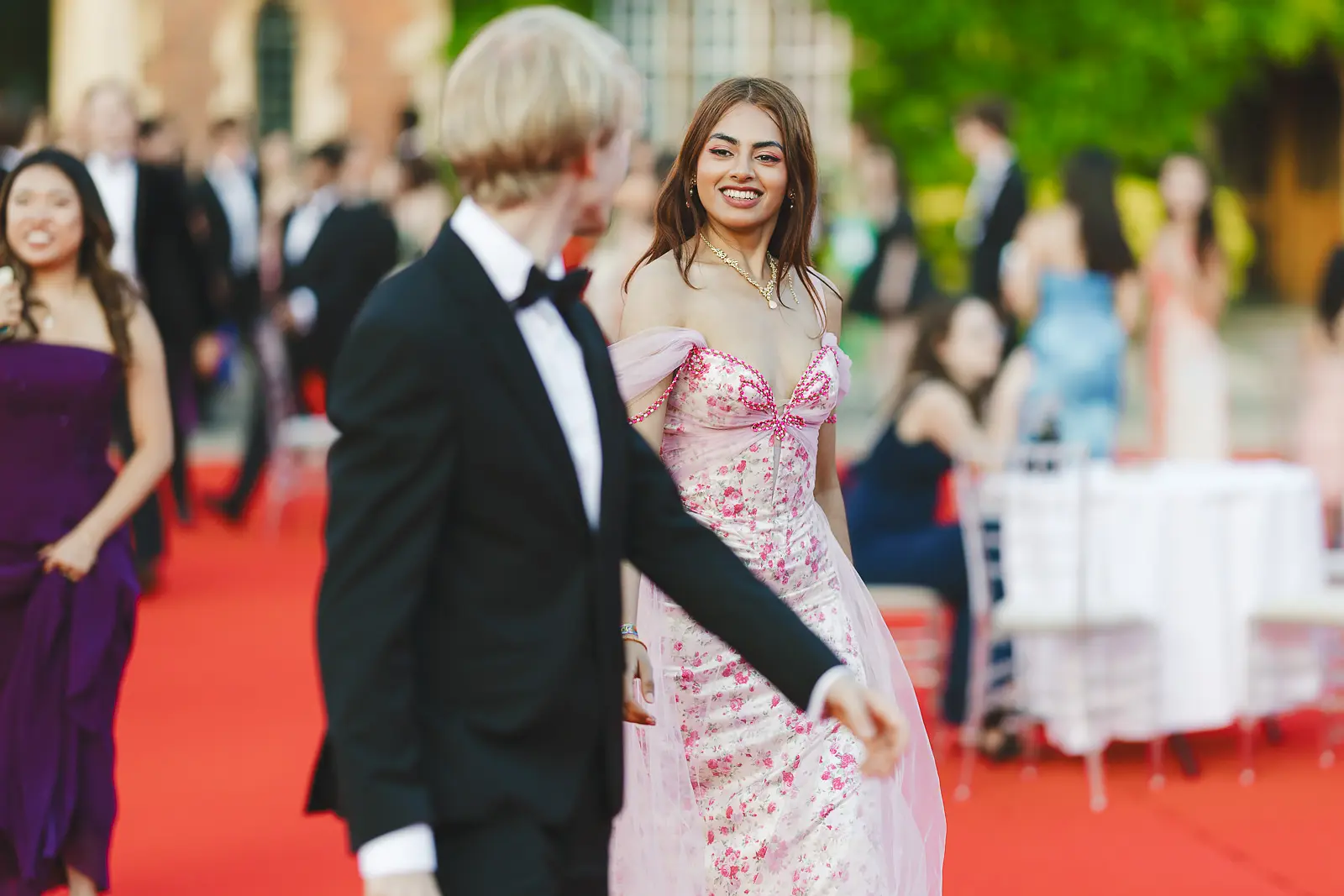 Guest in a pink floral off-shoulder ball gown smiling on the red carpet at a formal Cambridge ball