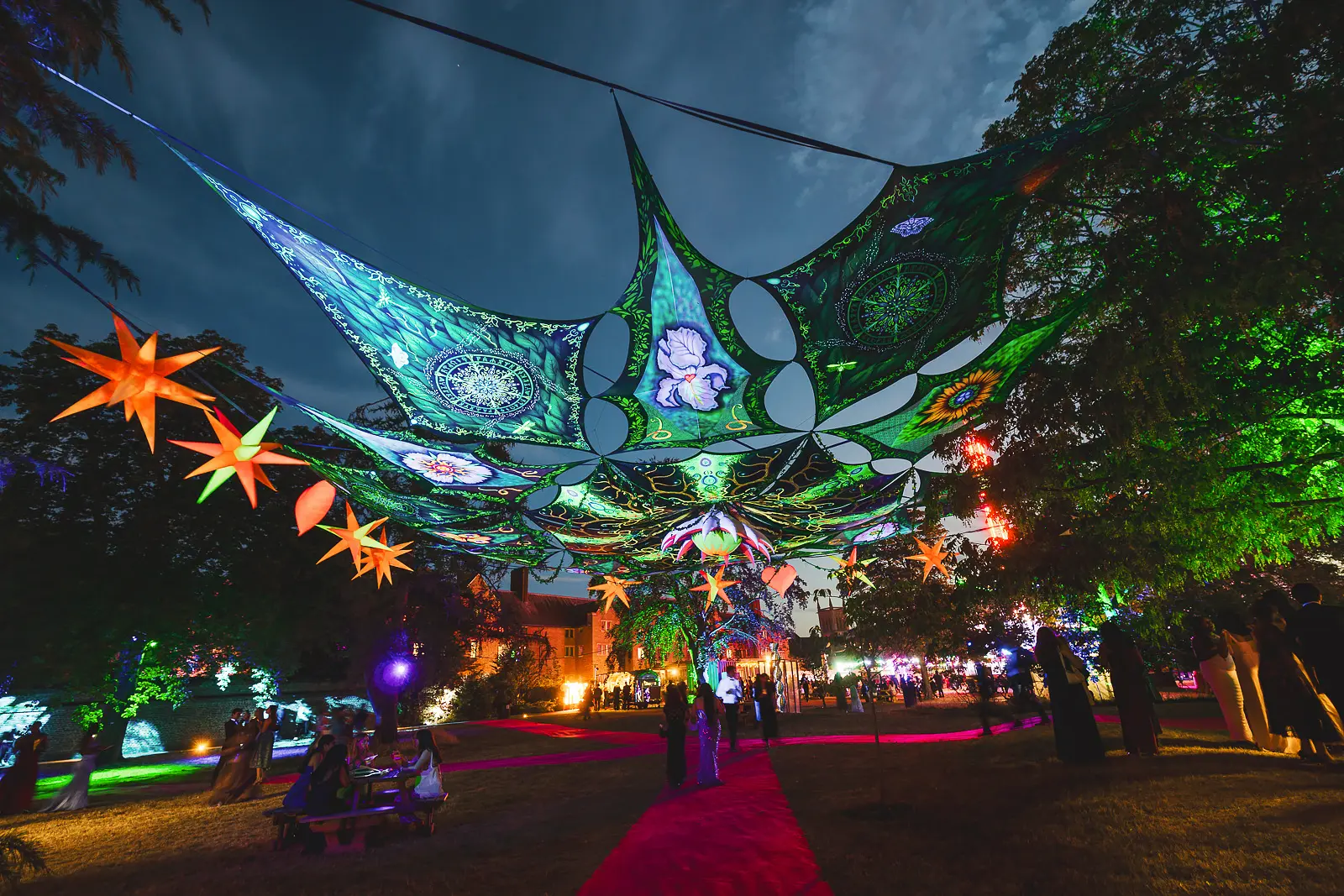 Psychedelic illuminated fabric canopy installation with star lanterns stretched between trees at a surrealist-themed summer ball