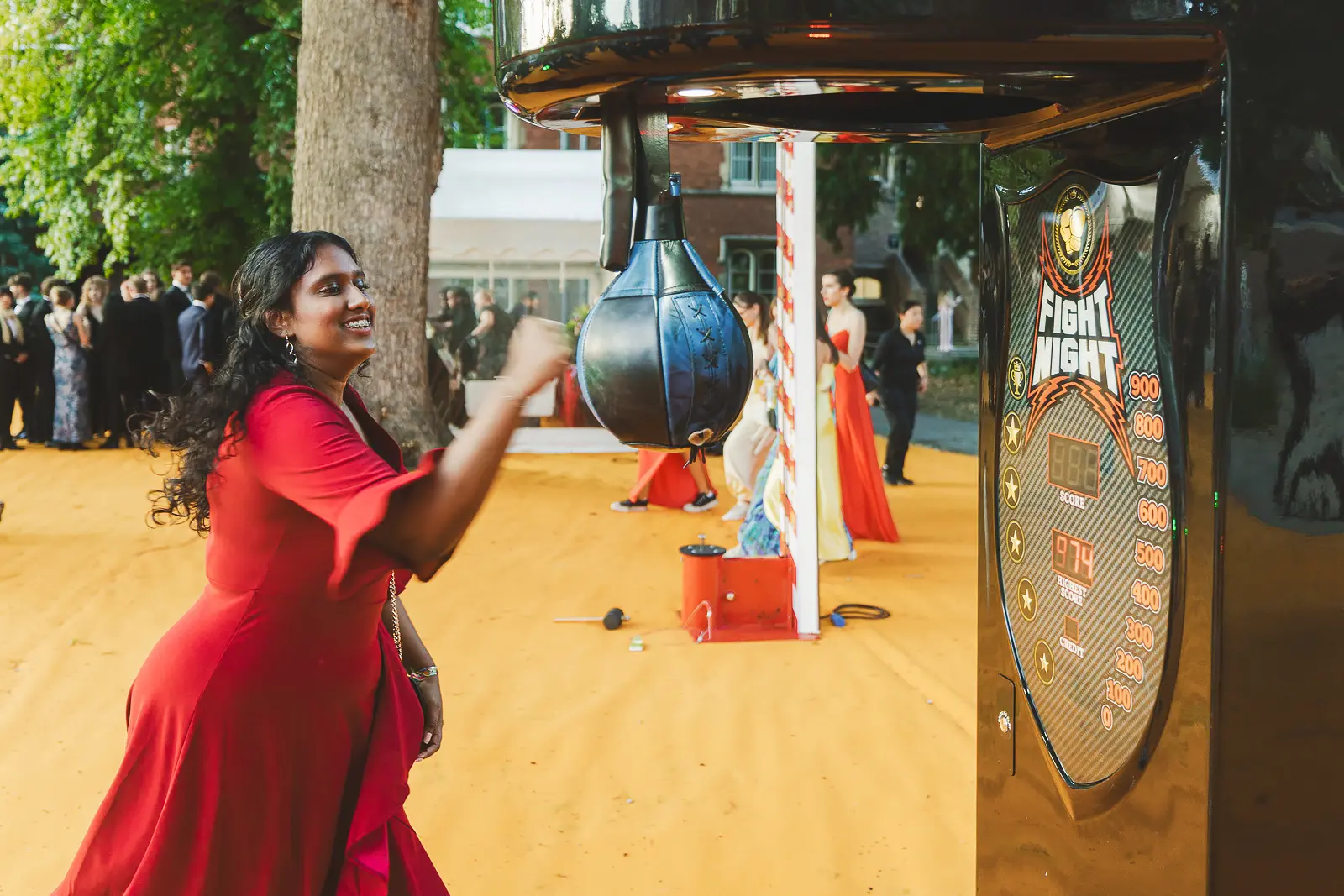 Guest in a red dress taking on a punch bag arcade machine at a May Ball entertainment attraction