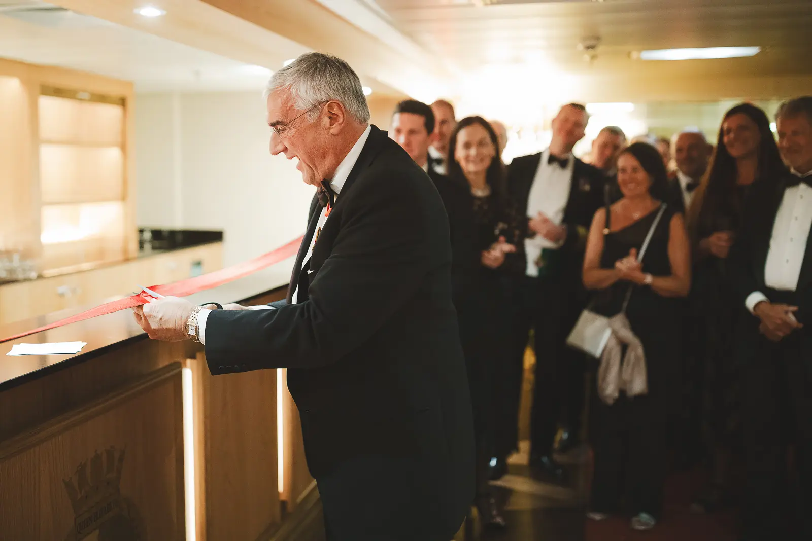 Civilian dignitary cutting red ribbon at formal launch ceremony, smiling crowd of black-tie guests watching in warm-lit ship interior