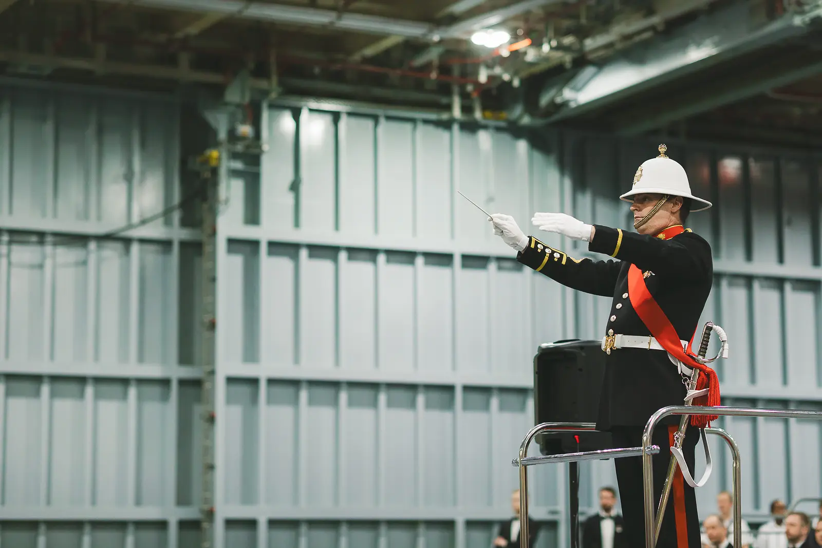 Royal Marines Band conductor in ceremonial dress with white pith helmet raising baton on podium inside aircraft carrier hangar