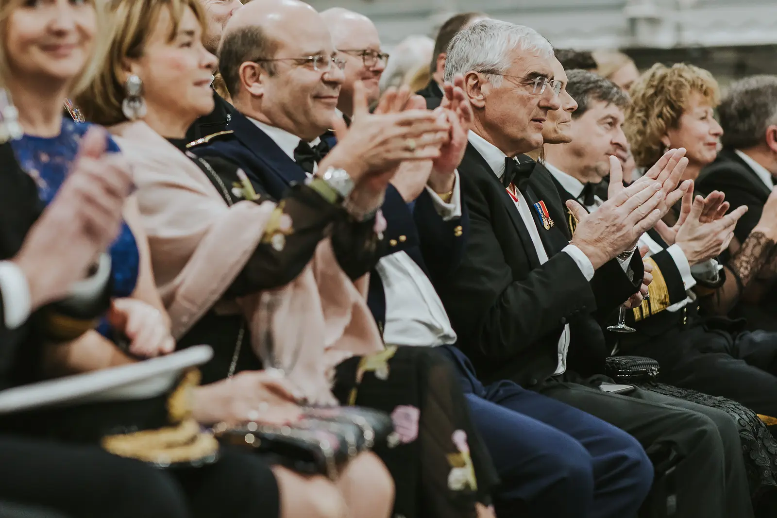 Row of seated guests in formal attire applauding enthusiastically during band performance, military officers and civilians together