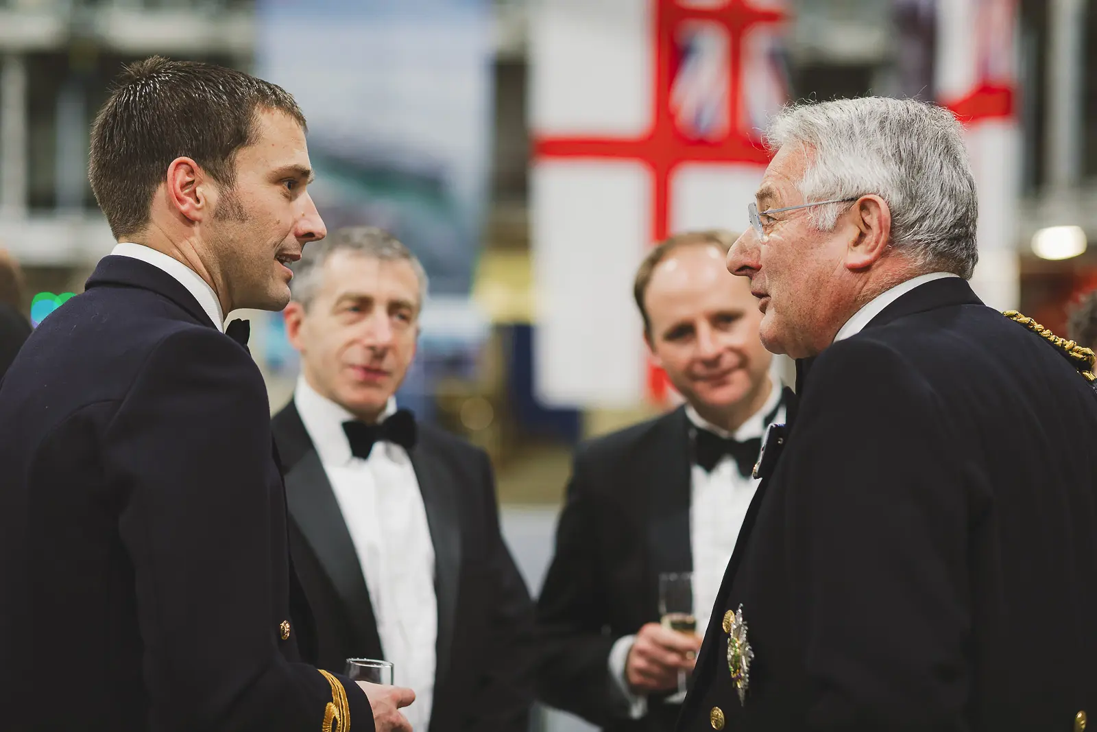 Senior officer with chain of office in conversation with junior officer at formal gala, civilian guests and flags in background
