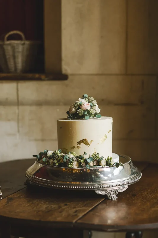 Single tier wedding cake with gold leaf detail and fresh flowers on a silver stand in the tithe barn at Hinton St Mary Estate